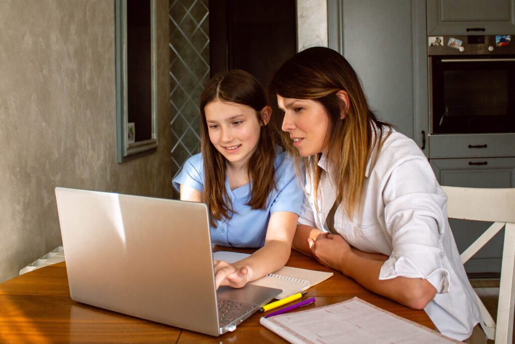 Daughter showing mom something exciting on internet on laptop screen sitting at table in kitchen. Happy parenthood and motherhood. Happy family concept